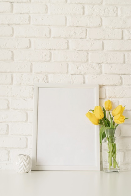 Boeket van gele tulpen in een glazen vaas en een leeg fotolijstje op een witte bakstenen muur achtergrond. Mock-up ontwerp