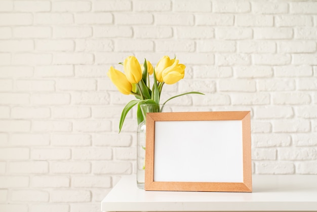 Boeket van gele tulpen in een glazen vaas en een leeg fotolijstje op een witte bakstenen muur achtergrond. Mock-up ontwerp