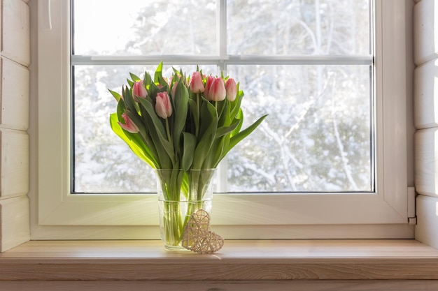 Boeket van mooie roze tulpen op een houten vensterbank Wit raam in houten huis in Scandinavische stijl met uitzicht op de tuin dennenbos winterweer Lente concept Happy 8 maart Womens Day