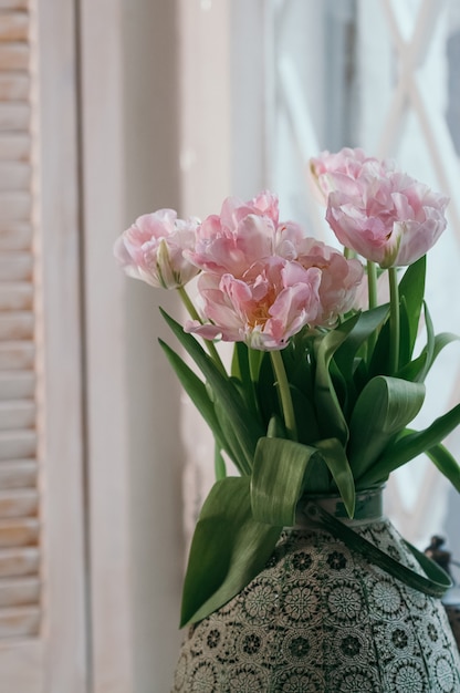 boeket van roze pioen tulpen in oude grote vaas op vensterbank licht houten jaloezie op achtergrond