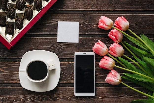 Boeket van roze tulpenbloemen met een kopje koffie, een doos chocolaatjes en een smartphone