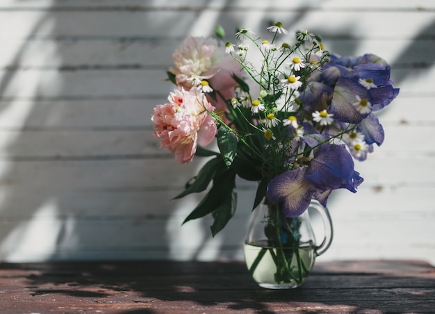 Boeket van witte pioenrozen, kamilles en irisbloemen in glazen vaas. Zomer achtergrond. Getinte foto.