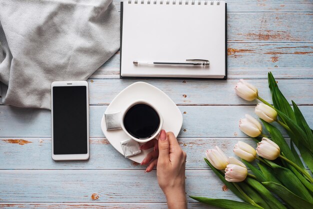 Boeket van witte tulpen met een kopje koffie in de handen van vrouwen, een smartphone en een leeg notitieboekje