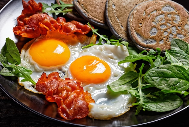 Boekweitpannenkoek met gebakken eieren, gebakken spek en groene salade geserveerd op een zwarte plaat op een houten tafel, ochtendontbijt, horizontale weergave van bovenaf, close-up