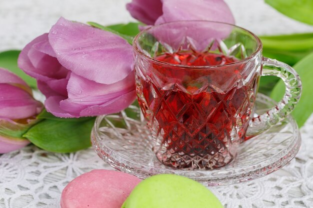 Bohemian crystal transparent cup of karkadeh red tea with cookies macaroons and perple tulips on a beautiful crochet tablecloth