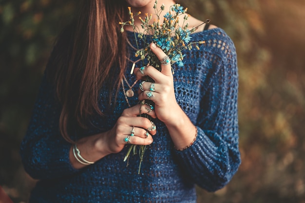 Boho chique vrouw in gebreide blauwe trui en het dragen van zilveren ringen met turquoise steen met boeket van wilde bloemen in handen in de herfst bos buiten in de herfst