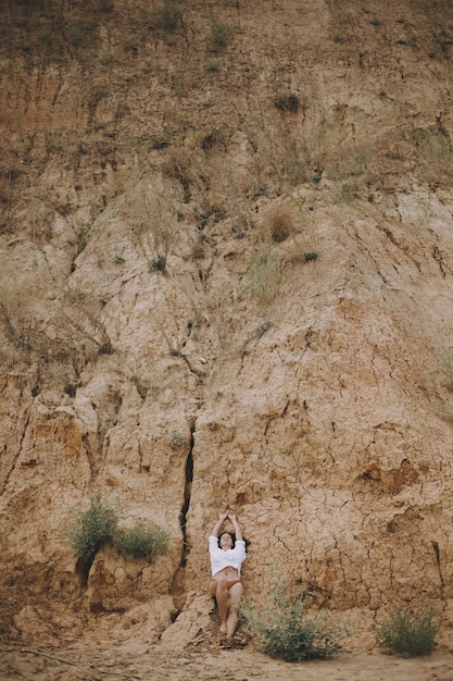 Boho girl in white shirt lying on sandy cliff on beach creative
aesthetic image carefree young woman relaxing on sand seashore
natural concept connection to nature