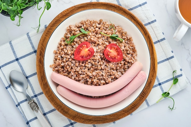 Boiled buckwheat porridge, meat sausages, tomato and microgreens of peas in a white plate in stone grey background. Fun food art idea healthy lunch for kids. Top view.