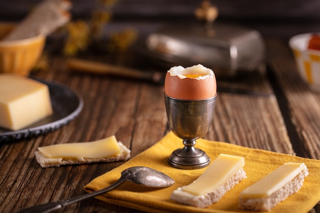 Boiled egg in a silver eggcup with bread and cheese on a wooden table