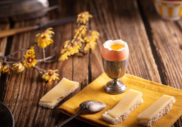 Boiled egg in a silver eggcup with bread and cheese on a wooden table