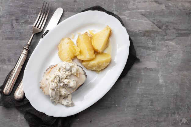 Вареная рыба с соусом тартар и отварным картофелем на белом блюде на керамическом столе