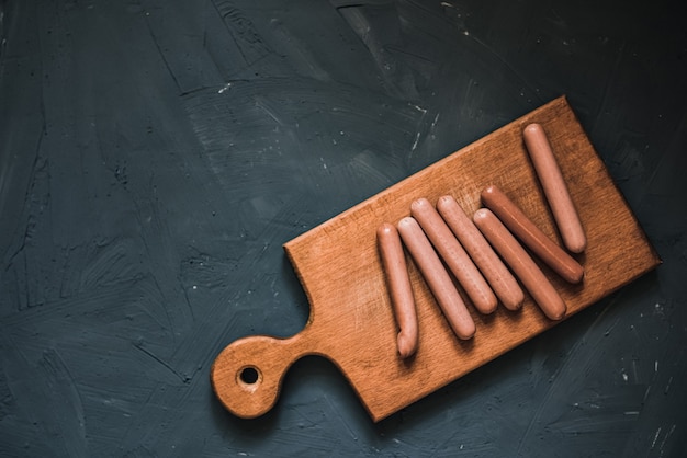 Boiled fried sausages sausages lie on a wooden kitchen board scratched against a dark concrete background.