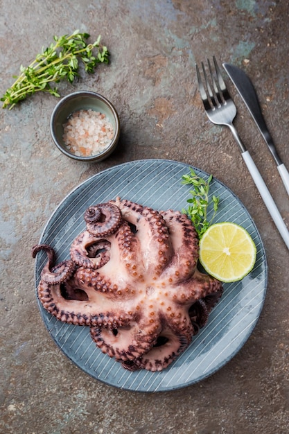 Boiled octopus ready for serve with lemon and thyme on plate over stone background, top view