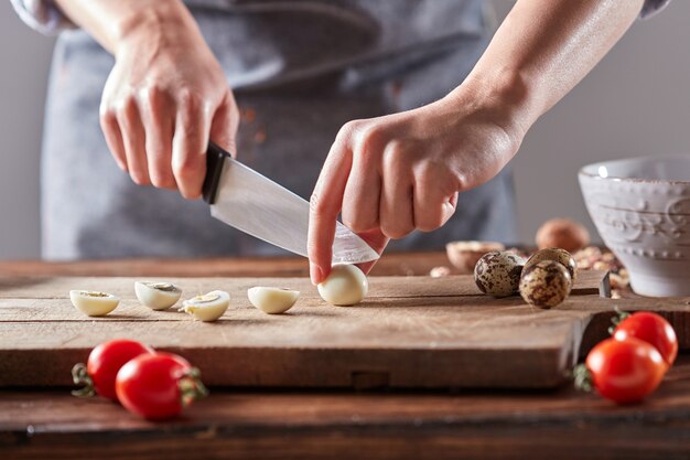ウズラのゆで卵は、トマトとほうれん草のテーブルの上にある木の板で女性の手を切ります。ステップバイステップの準備