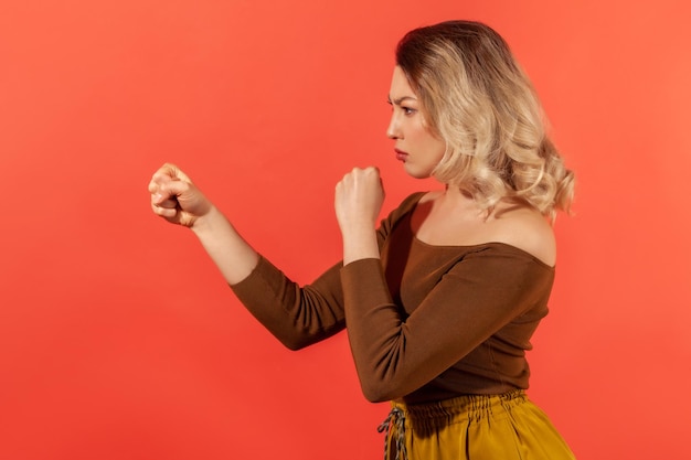 Bokser. Zijaanzicht portret van jonge vrouw met blond haar die beide vuisten stoot om te vechten en aandachtig naar de tegenstander te staren, ze uiting geven aan vertrouwen en macht Indoor geïsoleerd op rode achtergrond