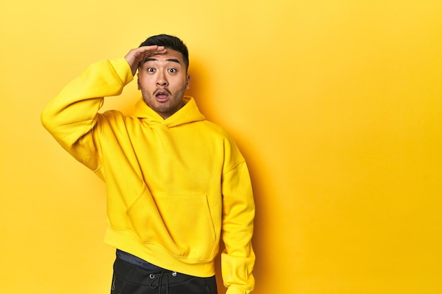 Bold Asian man in yellow hoodie on yellow studio shouts loud keeps eyes opened and hands tense