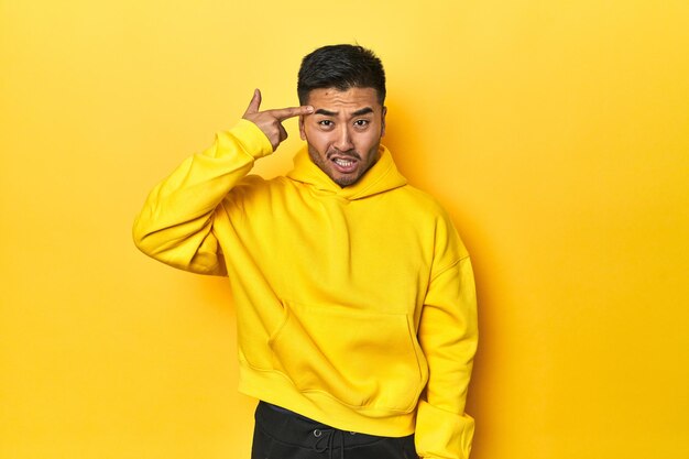 Bold Asian man in yellow hoodie on yellow studio showing a disappointment gesture with forefinger