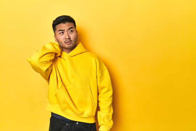 Photo bold asian man in yellow hoodie on yellow studio touching back of head thinking and making a choice