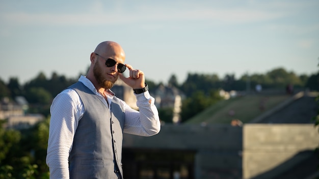 Photo bold guy with a stylish beard and sunglasses on a blurred city surface during sunset