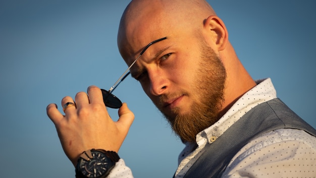 Bold Guy with a stylish beard and sunglasses on a sky surface during sunset