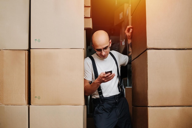 Bold tired male warehouse worker leaning on stack of boxes looking at his phone