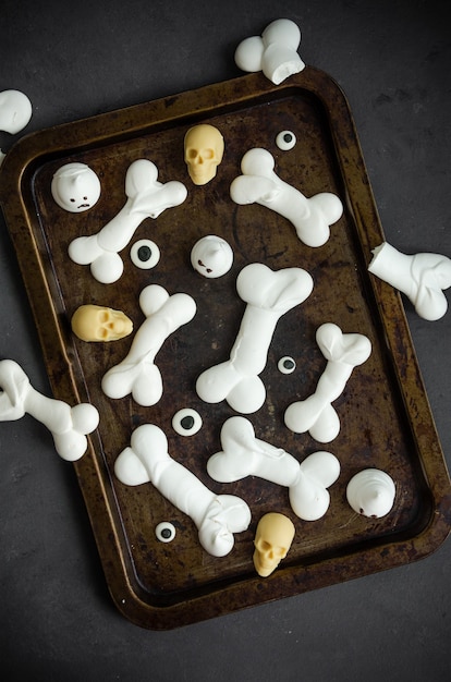 Bones Halloween Meringues with ghosts, chocolate skulls and eyes on an old baking sheet on a dark background. Food idea for halloween party. Top view, copy space.