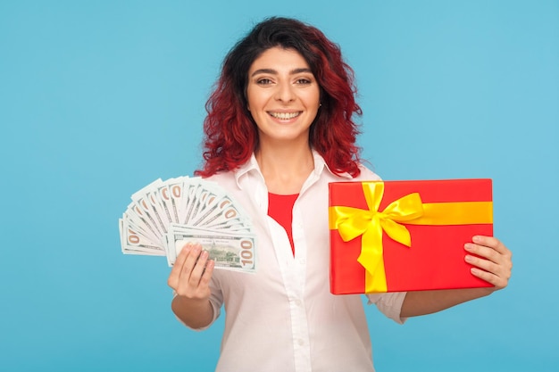 Bonus and cashback holiday discount Portrait of delighted woman with fancy red hair holding gift box and money enjoying presents from store shopping indoor studio shot isolated on blue background