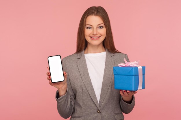 Bonus for mobile user online shopping application Portrait of happy elegant businesswoman in suit jacket holding cellphone and gift box smiling to camera studio shot isolated on pink background