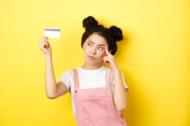 Boodschappen doen. Nadenkend trendy meisje met zomermake-up, peinzend kijken naar plastic creditcard, staande op geel.