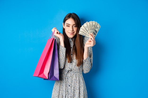 Boodschappen doen. Opgewonden aantrekkelijke vrouw die cadeaus voor zichzelf koopt, dollarbiljetten en kleurzakken uit de winkel toont, glimlachend tevreden, staande op blauw.