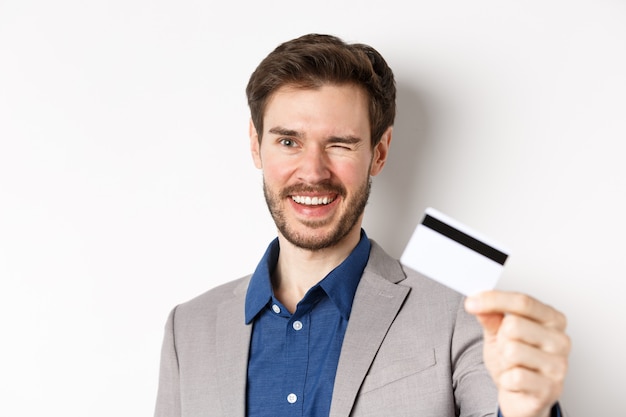 Boodschappen doen. Vrolijke bebaarde man in pak knipogen en glimlachen, plastic creditcard tonen op camera, witte achtergrond.