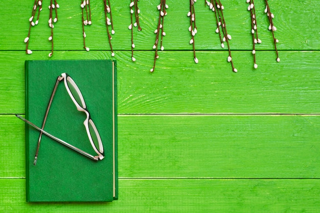 A book in a hard green cover on a green wooden background with glasses and willow branche