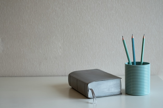 A book and pastel pencils in the blue glass on the desk at gray wall background