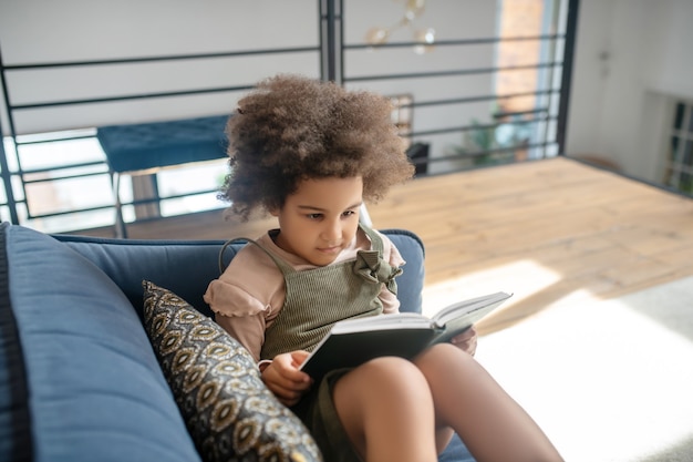 Book reading. Serious little african american girl with dark curly hair carefully looking at book on sofa in bright room