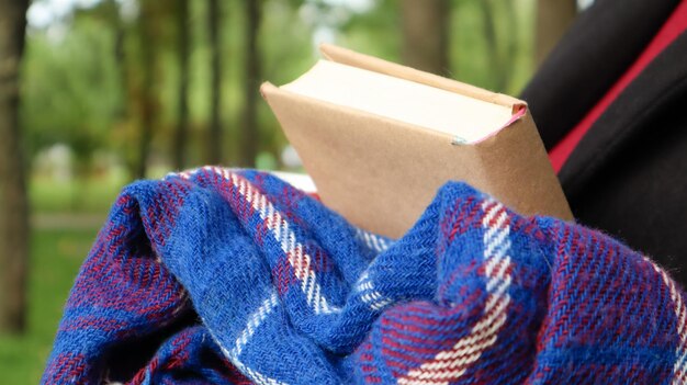 A book with a blue checkered woolen blanket or plaid in the hands of a woman wearing a sweater and black coat in the park. Warm and sunny weather. Soft, cozy photograph. Close-up.