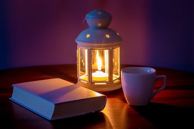 A book with a white cover near the lantern and a cup of coffee in the evening