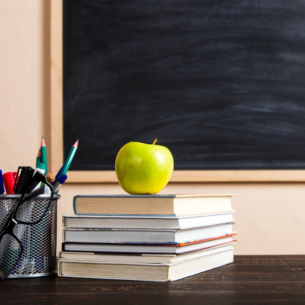 Books, apple, pens, pencils and glasses on a wooden table, against a chalk board.