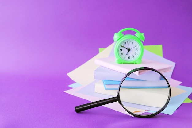 Books magnifying glass alarm clock and notepads pastel colored\
paper purple background