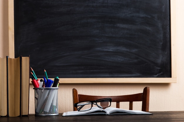 Books, pens, pencils and glasses on a wooden table, against a chalk board.