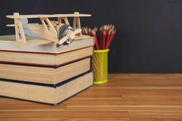 積み重ねられた本と鉛筆ホルダーにはたくさんの赤い木製の鉛筆がテーブルに置かれている飛行機のモデル背景は黒板です学校に戻るコンセプト