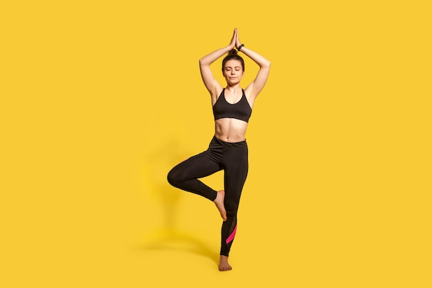 Boom pose atletische geconcentreerde vrouw in strakke sportkleding beoefenen van yoga mediteren met gesloten ogen doen Vrksasana oefening op één been handen omhoog volledige lengte studio shot sport trainingen geïsoleerd