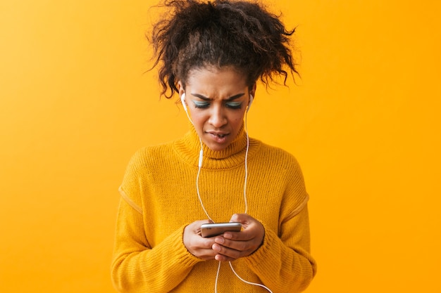 Foto boos afrikaanse vrouw, gekleed in trui staan geïsoleerd, luisteren naar muziek met koptelefoon, mobiele telefoon vast te houden