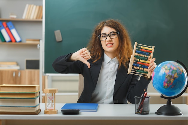 boos, duimen omlaag, jonge vrouwelijke leraar die een bril draagt en een telraam vasthoudt dat aan het bureau zit met schoolhulpmiddelen in de klas