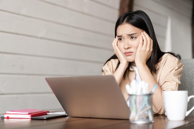 Boos en humeurig aziatisch meisje zit thuis in de buurt van laptop en kijkt teleurgesteld moe van het studeren op afstand...