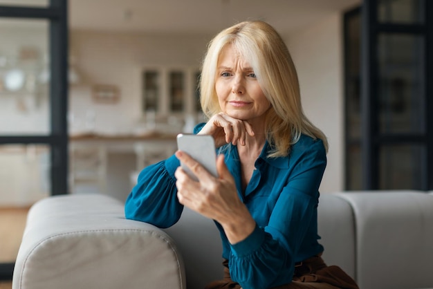 Boos europese rijpe vrouw zittend op de bank thuis met mobiele telefoon kijkend naar gadgetscherm