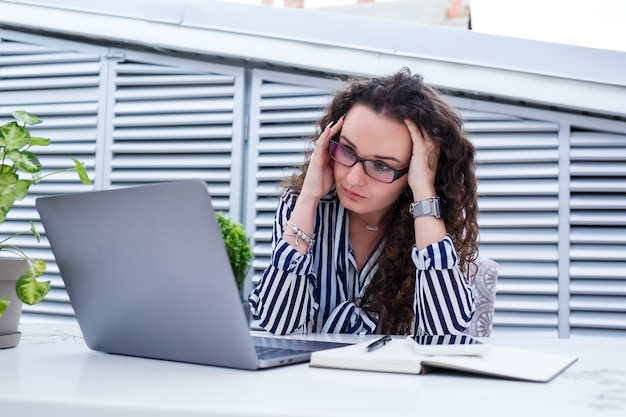 Boos freelancer vrouw in modieuze vrijetijdskleding, werken met een laptop en aantekeningen maken, glimlachen en kijken naar het terras van het café. zakelijke problemen en hoofdpijn