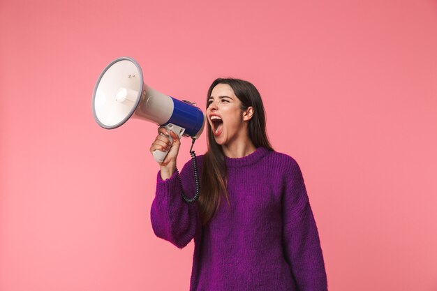 Boos jong meisje die sweater dragen die zich geïsoleerd over roze bevinden, die in een spreker schreeuwen