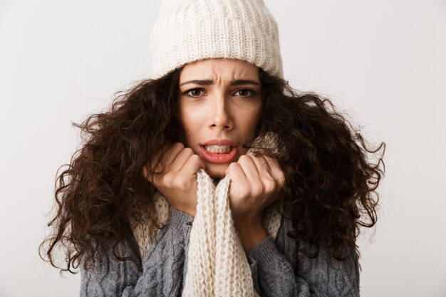 Boos jonge vrouw die de wintersjaal draagt ?? die zich geïsoleerd over witte muur bevindt, rilt