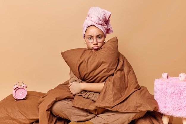 Boos jonge vrouw in bril omarmt kussen gewikkeld in deken heeft handdoek op hoofd poses in bed wil niet wakker worden geïsoleerd over bruine achtergrond. Ochtendtijd en huiselijke sfeer.