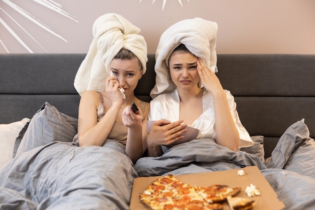 Boos meisjes liggend op bed, tv of film kijken en huilen. Jonge Europese vrouwen met ingepakte badhanddoeken op hoofden. Concept van meisjesfeest thuis. Interieur van slaapkamer in modern appartement